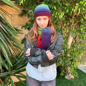 May include: A rainbow-coloured knitted beanie hat and scarf set. The beanie and scarf feature a gradient of colours, including purple, blue, green, and red. The set is worn with a black jacket, white shirt, and grey trousers. Green foliage is in the background.