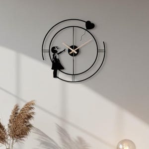 May include: Black metal wall clock with a silhouette of a girl reaching for a heart-shaped balloon. The clock has a circular design with beige clock hands. A decorative piece for any room.