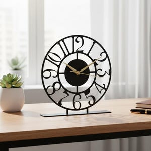 Może przedstawiać: Czarny, metalowy zegar biurkowy z okrągłą tarczą i wyciętymi cyframi. Zegar ma złote wskazówki i stoi na prostokątnej podstawie. Zegar jest wystawiony na drewnianym biurku.
