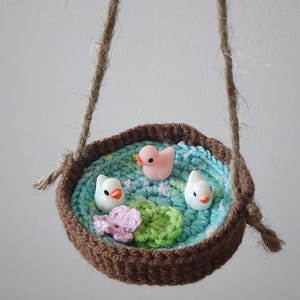 Peut inclure: Une scène de bassin au crochet faite à la main avec trois figurines de canards, une fleur rose et des nénuphars verts. Le bassin est dans un panier en crochet marron avec une poignée en corde de jute. La scène est suspendue.