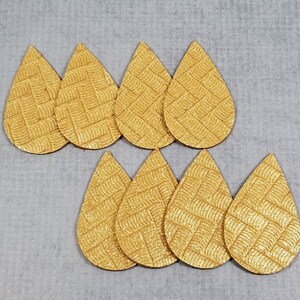 May include: A collection of eight gold-colored, teardrop-shaped leather earring cutouts. Each cutout has a textured, woven pattern. The cutouts are arranged on a light gray surface.
