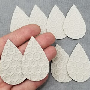 May include: A collection of light gray teardrop-shaped leather earring blanks. The blanks have a textured honeycomb pattern. Several are arranged on a gray surface, with some held in a hand.