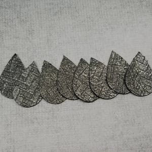 May include: A collection of seven silver teardrop-shaped earrings with a textured, woven pattern. The earrings are arranged in a row against a light gray background, showcasing their metallic sheen and unique design.