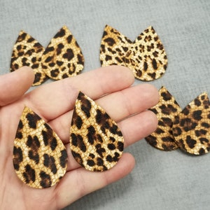 K&ouml;nnte beinhalten: Tropfenf&ouml;rmige Ohrringe mit Leopardenmuster. Die Ohrringe sind braun und beige mit dunkelbraunen Flecken. Die Ohrringe werden in einer Hand vor grauem Hintergrund gezeigt. Die Ohrringe sind aus Leder.
