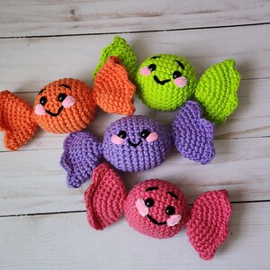 May include: Crocheted candy toys in orange, green, purple, and pink. Each candy has a smiling face with black eyes and a pink blush. The candies are arranged on a light wood-grain surface.