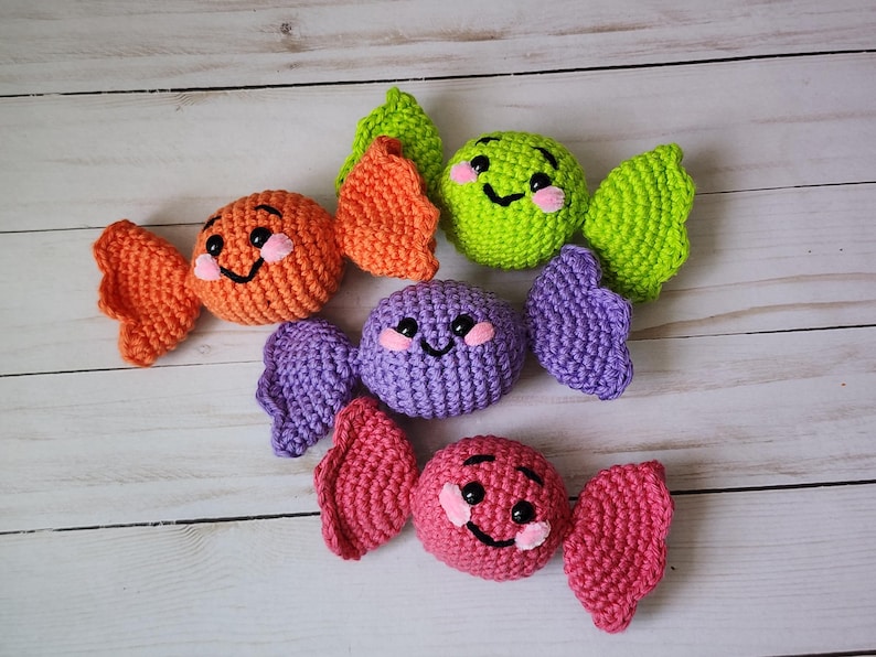 May include: Crocheted candy toys in orange, green, purple, and pink. Each candy has a smiling face with black eyes and a pink blush. The candies are arranged on a light wood-grain surface.
