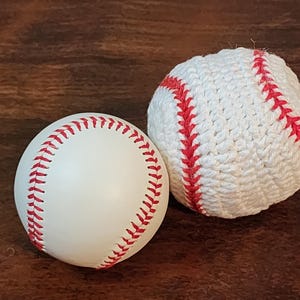 May include: Two baseballs on a brown surface. One is a standard white baseball with red stitching. The other is a white, crocheted baseball with red stitching. The baseballs are side by side.