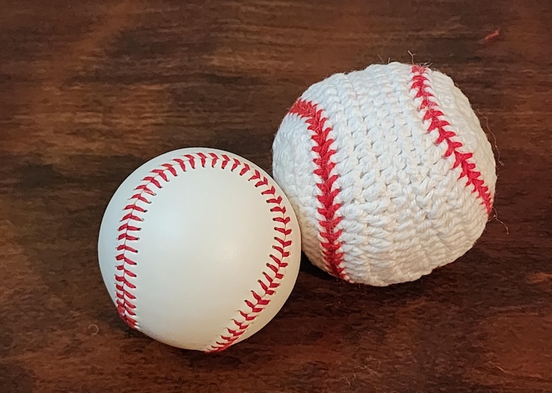 May include: Two baseballs on a brown surface. One is a standard white baseball with red stitching. The other is a white, crocheted baseball with red stitching. The baseballs are side by side.