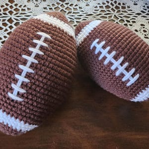 Peut inclure: Deux ballons de football américains crochetés à la main, marron avec des coutures et des rayures blanches. Les ballons sont posés sur une surface marron foncé, avec une nappe en dentelle blanche en arrière-plan. Jouet doux.