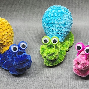 Può includere: Tre chiocciole all'uncinetto fatte a mano in colori vivaci. Una è blu con guscio giallo, un'altra è verde con guscio blu e la terza è rosa con guscio azzurro. Ognuna ha grandi occhi mobili.