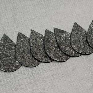 May include: A row of eight teardrop-shaped, textured, silver-colored pieces. The pieces are arranged in a curved line, with the points facing the same direction. The background is a light gray, textured surface.