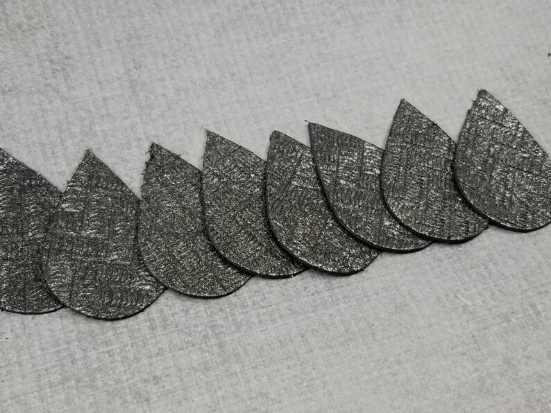 May include: A row of eight teardrop-shaped, textured, silver-colored pieces. The pieces are arranged in a curved line, with the points facing the same direction. The background is a light gray, textured surface.