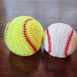 以下が含まれることがあります： 赤色のステッチが入った黄色のソフトボールと、赤色のステッチが入った白い野球の、2つの手編みのスポーツボール。手作りのボールが濃い木製の表面に置かれています。