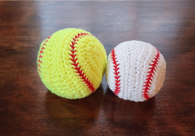 May include: Two crocheted sports balls: a yellow softball with red stitching and a white baseball with red stitching. The balls are handmade and sit on a dark wooden surface.