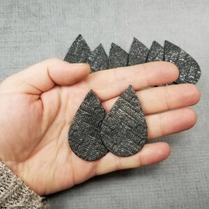 May include: Two dark gray, teardrop-shaped earrings with a textured pattern are held in a hand. Several similar earrings are visible in the background. The earrings have a metallic sheen and a unique design.