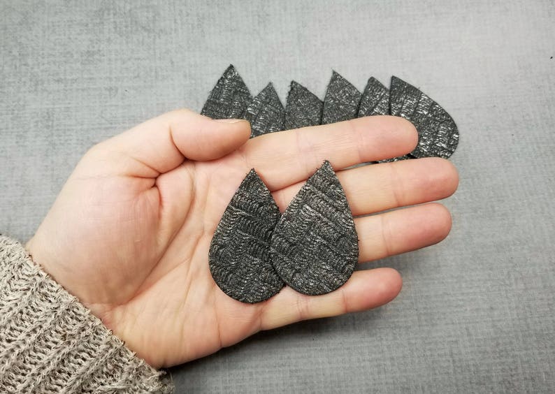 May include: Two dark gray, teardrop-shaped earrings with a textured pattern are held in a hand. Several similar earrings are visible in the background. The earrings have a metallic sheen and a unique design.