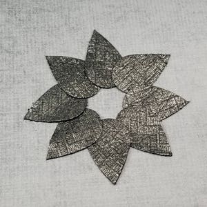 May include: A star-shaped arrangement of seven metallic gray leaf-shaped cutouts. Each cutout has a textured, woven pattern. The leaves are arranged in a circular pattern, with the tips pointing outwards. The background is a light gray, textured surface.