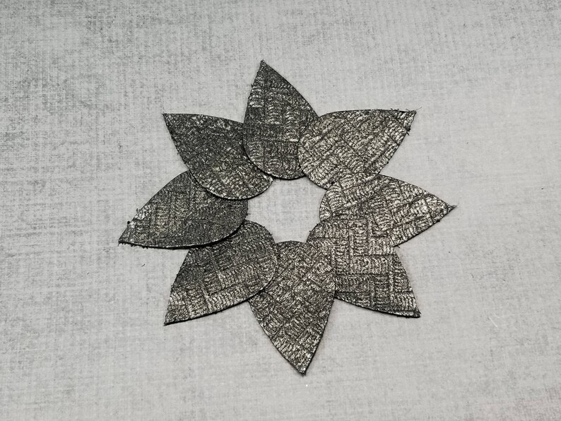 May include: A star-shaped arrangement of seven metallic gray leaf-shaped cutouts. Each cutout has a textured, woven pattern. The leaves are arranged in a circular pattern, with the tips pointing outwards. The background is a light gray, textured surface.