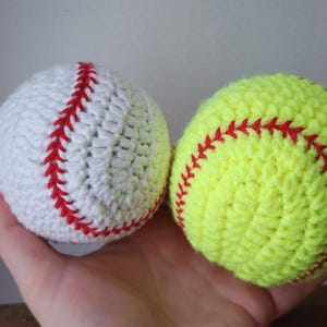 May include: Two crocheted sports balls: a white baseball with red stitching and a yellow softball with red stitching. The balls are handmade and appear soft, perfect for a sports-themed nursery or child's room.