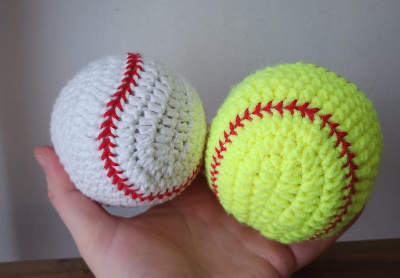 May include: Two crocheted sports balls: a white baseball with red stitching and a yellow softball with red stitching. The balls are handmade and appear soft, perfect for a sports-themed nursery or child's room.