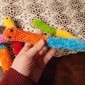 May include: Five colorful crocheted worm toys. The worms are made of soft, fuzzy yarn and have different colors, including pink, orange, yellow, green, and blue.