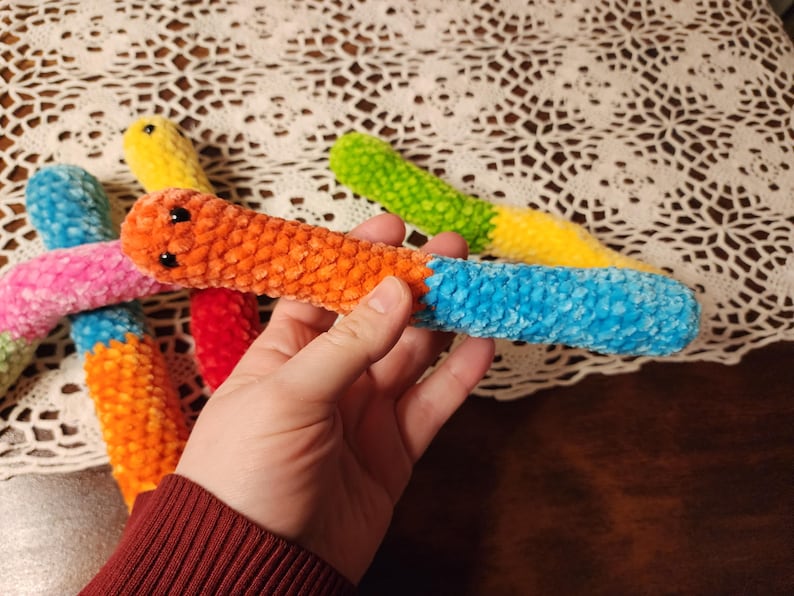 May include: Five colorful crocheted worm toys. The worms are made of soft, fuzzy yarn and have different colors, including pink, orange, yellow, green, and blue.