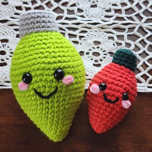 May include: Two handmade crochet Christmas light ornaments. One is lime green with a gray top, black eyes, and pink cheeks. The other is red with a green top, black eyes, and pink cheeks. Both have smiling faces.