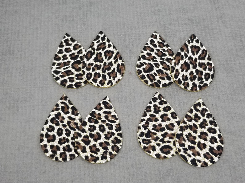 May include: Six teardrop-shaped leather earrings with a leopard print pattern. The earrings are white with black and brown spots.