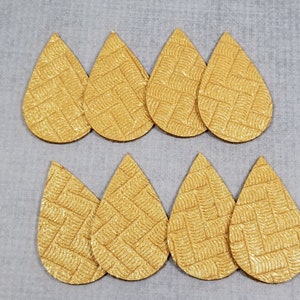 May include: Eight gold teardrop-shaped earring blanks with a textured woven pattern. The blanks are arranged in two rows of four, set against a light gray background. These are ideal for crafting earrings.