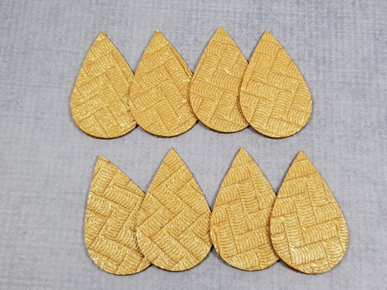 May include: Eight gold teardrop-shaped earring blanks with a textured woven pattern. The blanks are arranged in two rows of four, set against a light gray background. These are ideal for crafting earrings.