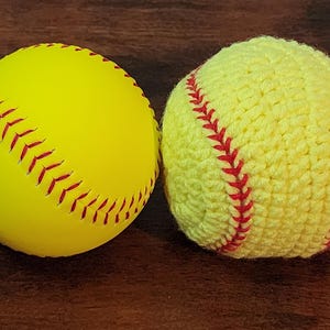 May include: Two softballs, one smooth and the other crocheted. The smooth softball is yellow with red stitching. The crocheted softball is also yellow with red stitching. Both are on a dark brown surface.