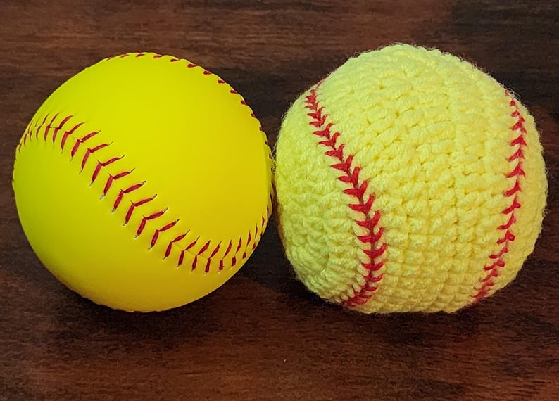May include: Two softballs, one smooth and the other crocheted. The smooth softball is yellow with red stitching. The crocheted softball is also yellow with red stitching. Both are on a dark brown surface.