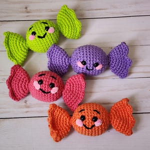 May include: Four colorful crocheted candies in green, purple, pink, and orange. Each candy has a smiling face with pink cheeks and black eyes. The candies are arranged on a light wood-grain surface.