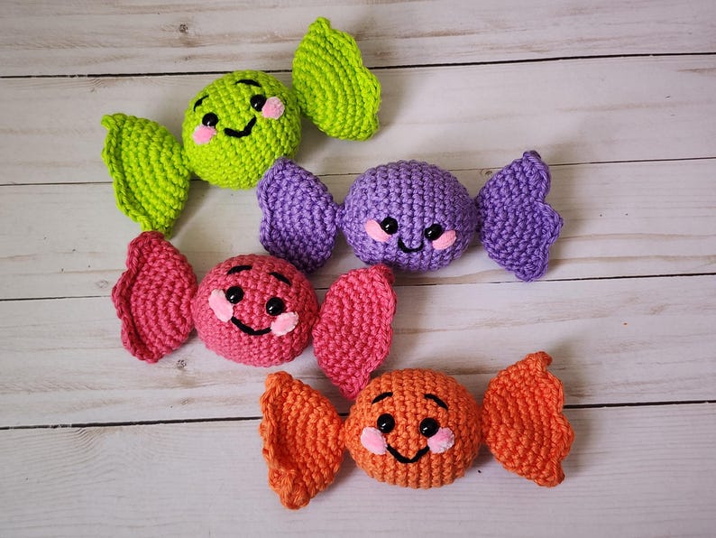 May include: Four colorful crocheted candies in green, purple, pink, and orange. Each candy has a smiling face with pink cheeks and black eyes. The candies are arranged on a light wood-grain surface.