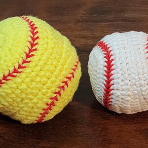 May include: Two crocheted sports balls: a yellow softball with red stitching and a white baseball with red stitching. The balls are handmade and sit on a dark brown surface. The softball is larger than the baseball.