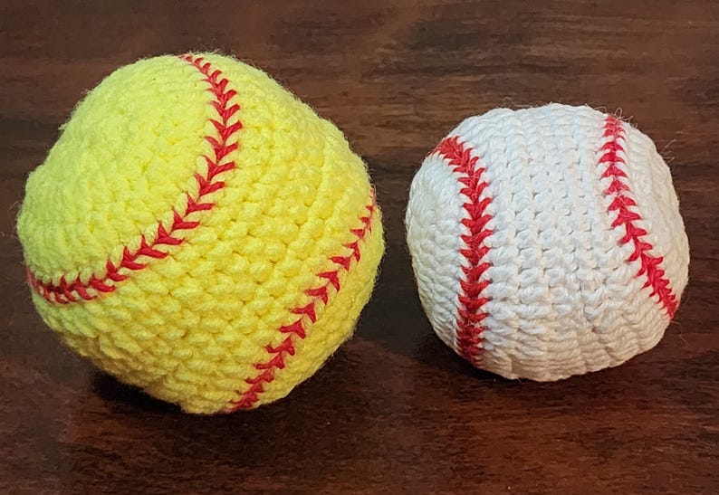 May include: Two crocheted sports balls: a yellow softball with red stitching and a white baseball with red stitching. The balls are handmade and sit on a dark brown surface. The softball is larger than the baseball.