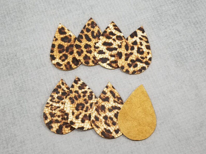 K&ouml;nnte beinhalten: Eine Sammlung tropfenf&ouml;rmiger Ohrring-Ausschnitte. Die nach vorne gerichteten Ausschnitte weisen ein tan- und braunes Leopardenmuster auf. Ein Ausschnitt wird mit der tanfarbenen Wildlederr&uuml;ckseite gezeigt. Die Ohrringe sind auf einer grauen Oberfl&auml;che angeordnet.