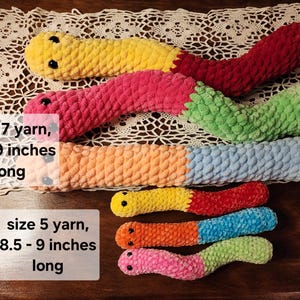 May include: Five crocheted worm plushies in various colors, including pink, green, blue, orange, and yellow. The worms are arranged in a row, with the largest worm at the top and the smallest worm at the bottom. The text "size 7 yarn, 18-19 inches long" is written below the largest worm, and the text "size 5 yarn, 8.5 - 9 inches long" is written below the smallest worm.