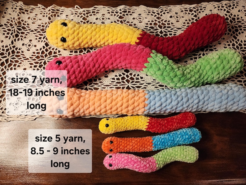 May include: Five crocheted worm plushies in various colors, including pink, green, blue, orange, and yellow. The worms are arranged in a row, with the largest worm at the top and the smallest worm at the bottom. The text "size 7 yarn, 18-19 inches long" is written below the largest worm, and the text "size 5 yarn, 8.5 - 9 inches long" is written below the smallest worm.