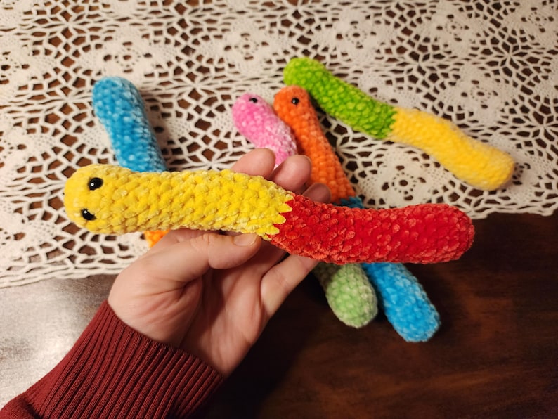 May include: A set of six colorful crocheted worm toys. The worms are made of soft, fuzzy yarn and have black eyes. The colors of the worms are blue, green, yellow, orange, pink, and red.