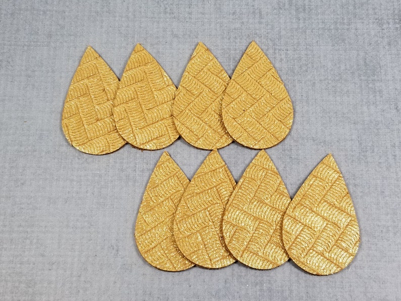 May include: A collection of eight gold-colored, teardrop-shaped leather earring cutouts. Each cutout has a textured, woven pattern. The cutouts are arranged on a light gray surface.