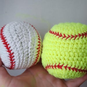 May include: Two handmade crochet sports balls: a white baseball with red stitching and a yellow softball with red stitching. The balls are held in a hand, showcasing the detailed crochet work and contrasting colors.