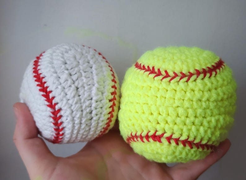 May include: Two handmade crochet sports balls: a white baseball with red stitching and a yellow softball with red stitching. The balls are held in a hand, showcasing the detailed crochet work and contrasting colors.