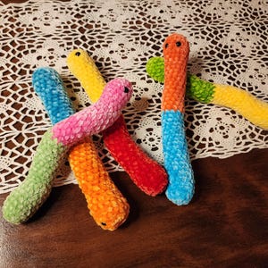 May include: Five colorful crocheted worm-shaped plush toys. The worms are green, yellow, orange, pink, and blue.