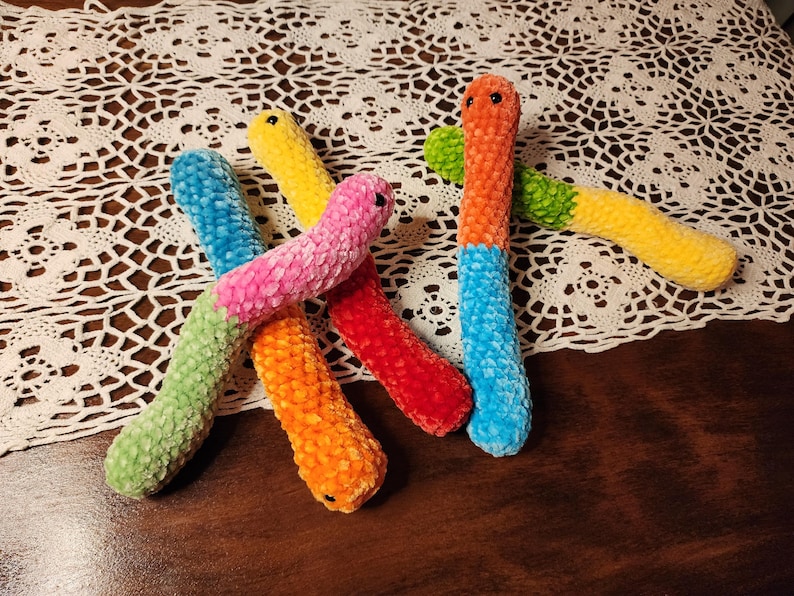 May include: Five colorful crocheted worm-shaped plush toys. The worms are green, yellow, orange, pink, and blue.