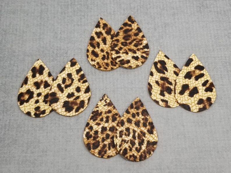 K&ouml;nnte beinhalten: Sechs Paar tropfenf&ouml;rmige Ohrring-Ausschnitte aus Kunstleder. Die Ausschnitte haben einen beigefarbenen Hintergrund mit braunen Leopardenflecken. Die Ohrringe sind auf einer grauen Oberfl&auml;che angeordnet.
