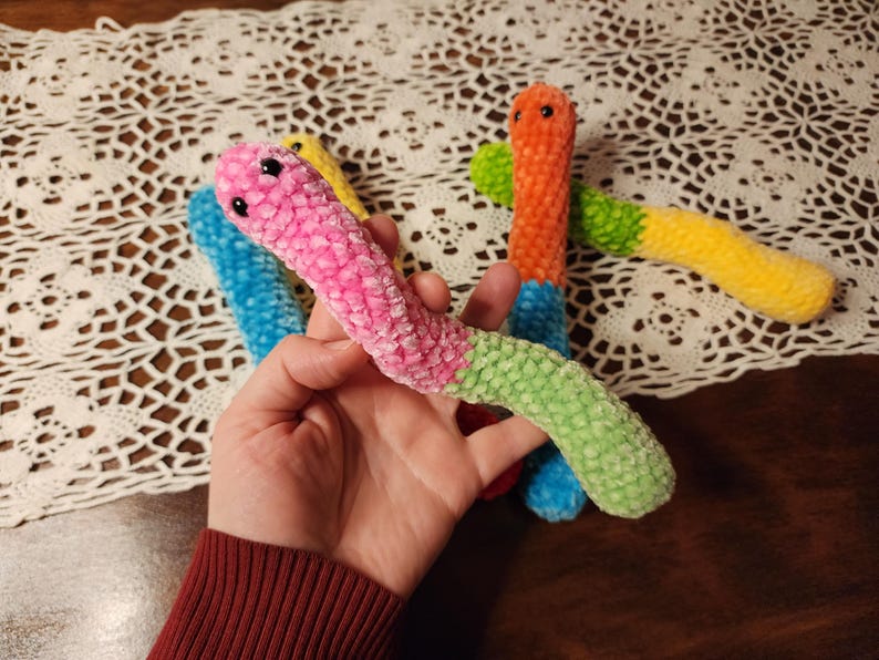 May include: Four colorful crocheted worm toys. The worms are pink, blue, green, and orange. They are all about 10 inches long and have a fuzzy texture.