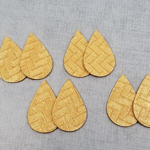May include: Six teardrop-shaped, textured, golden-yellow leather earring blanks. Each earring blank has a woven pattern. The blanks are arranged on a light gray surface.