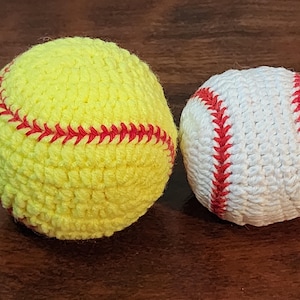 May include: Two handmade crochet sports balls. A yellow softball with red stitching and a white baseball with red stitching are displayed on a dark wood surface. The balls are round and textured, showcasing the detailed crochet work.