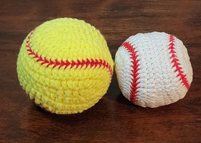 May include: Two handmade crochet sports balls. A yellow softball with red stitching and a white baseball with red stitching are displayed on a dark wood surface. The balls are round and textured, showcasing the detailed crochet work.